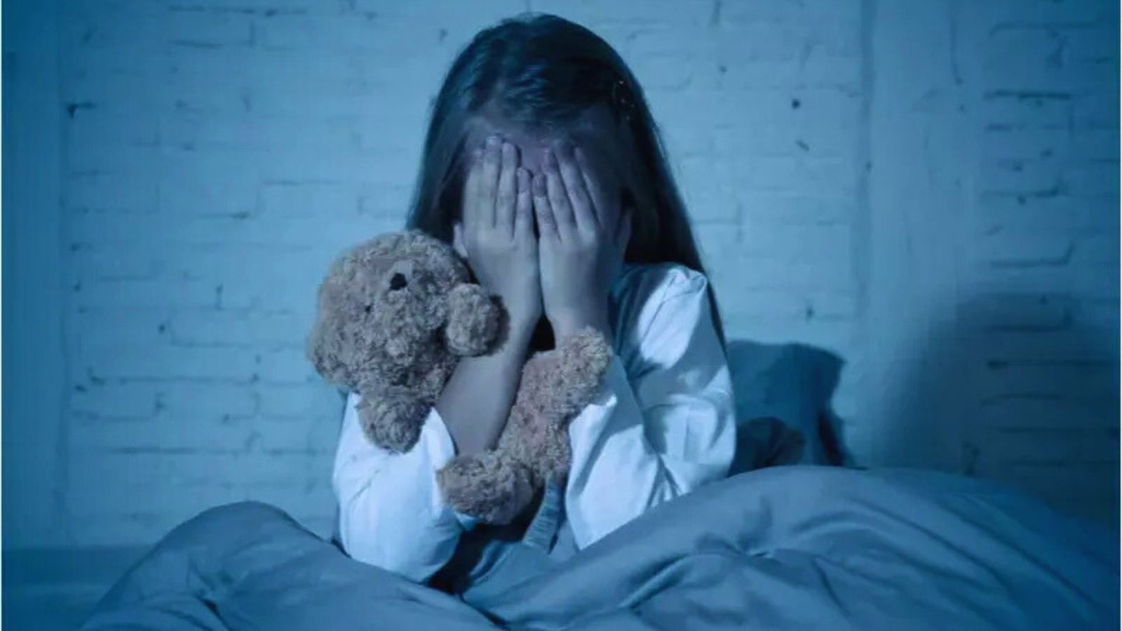In bed, a young girl holds a teddy bear tightly, finding solace from her fear of the dark.

