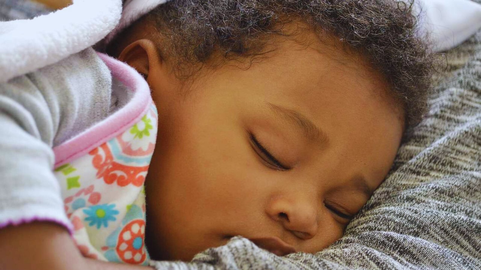 A baby peacefully sleeps on a blanket, resting her head on her mother's shoulder during a nap transition.
