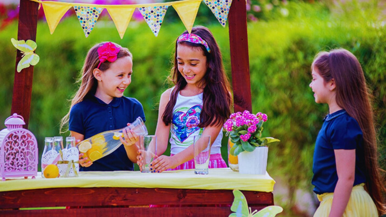 . Three little girls stand around a table, excitedly selling lemonade at their colorful lemonade stand.
