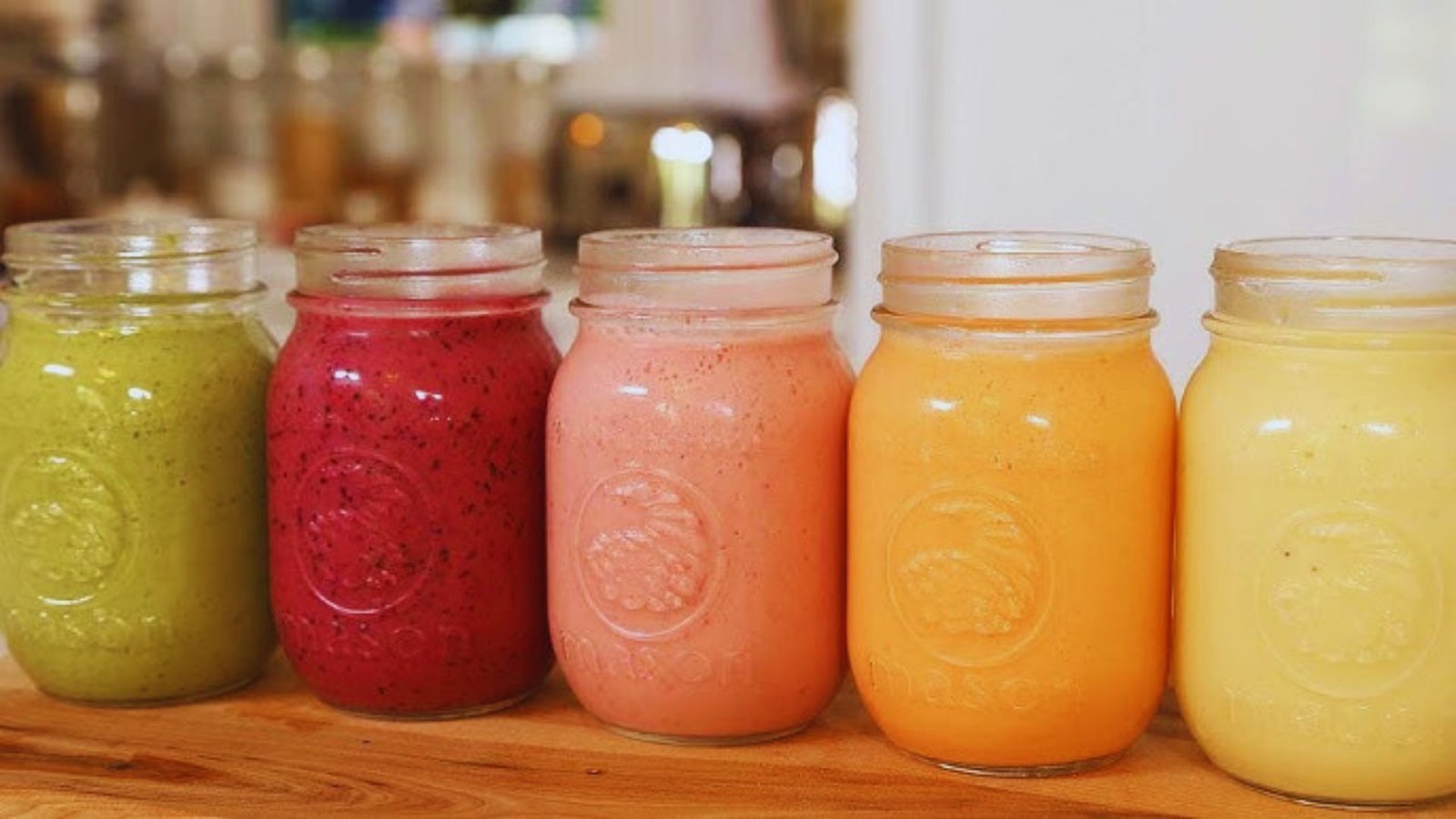 Five colorful smoothies in jars arranged neatly on a table, showcasing a variety of vibrant hues.
