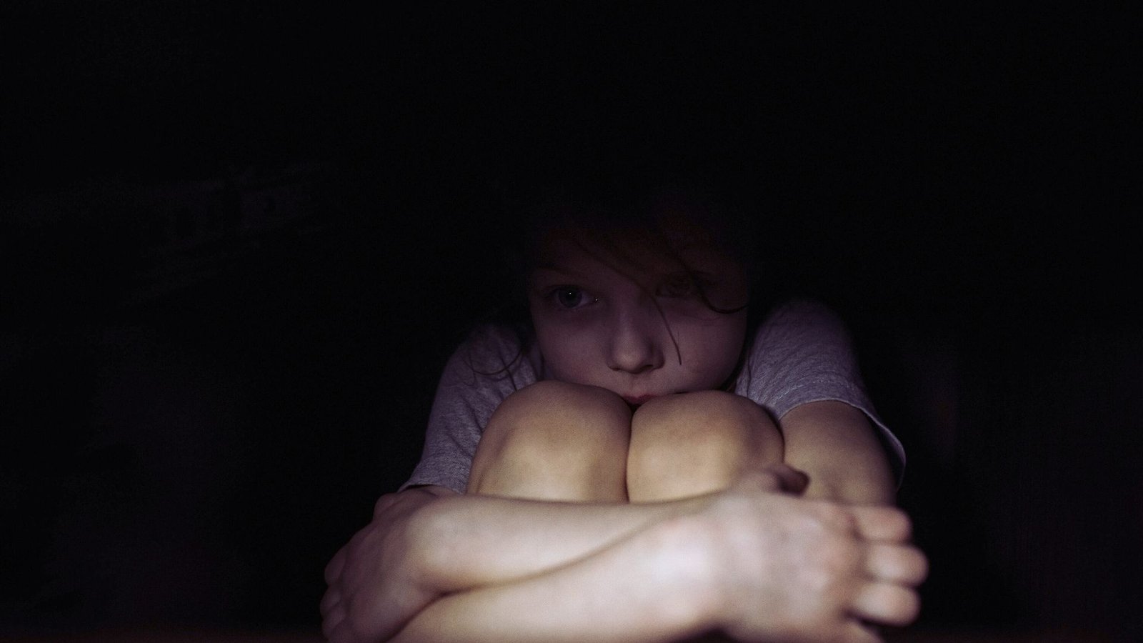 A young girl sits in the dark, hands on her knees, reflecting a common childhood fear of the dark.
