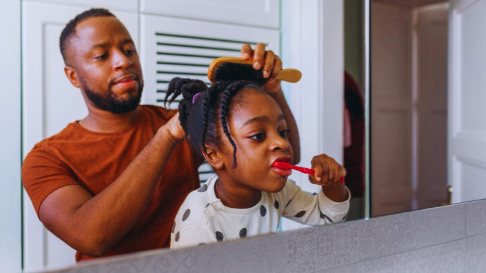 A father helps his daughter brush her teeth in a bright bathroom, promoting good dental hygiene in their daily routine.
