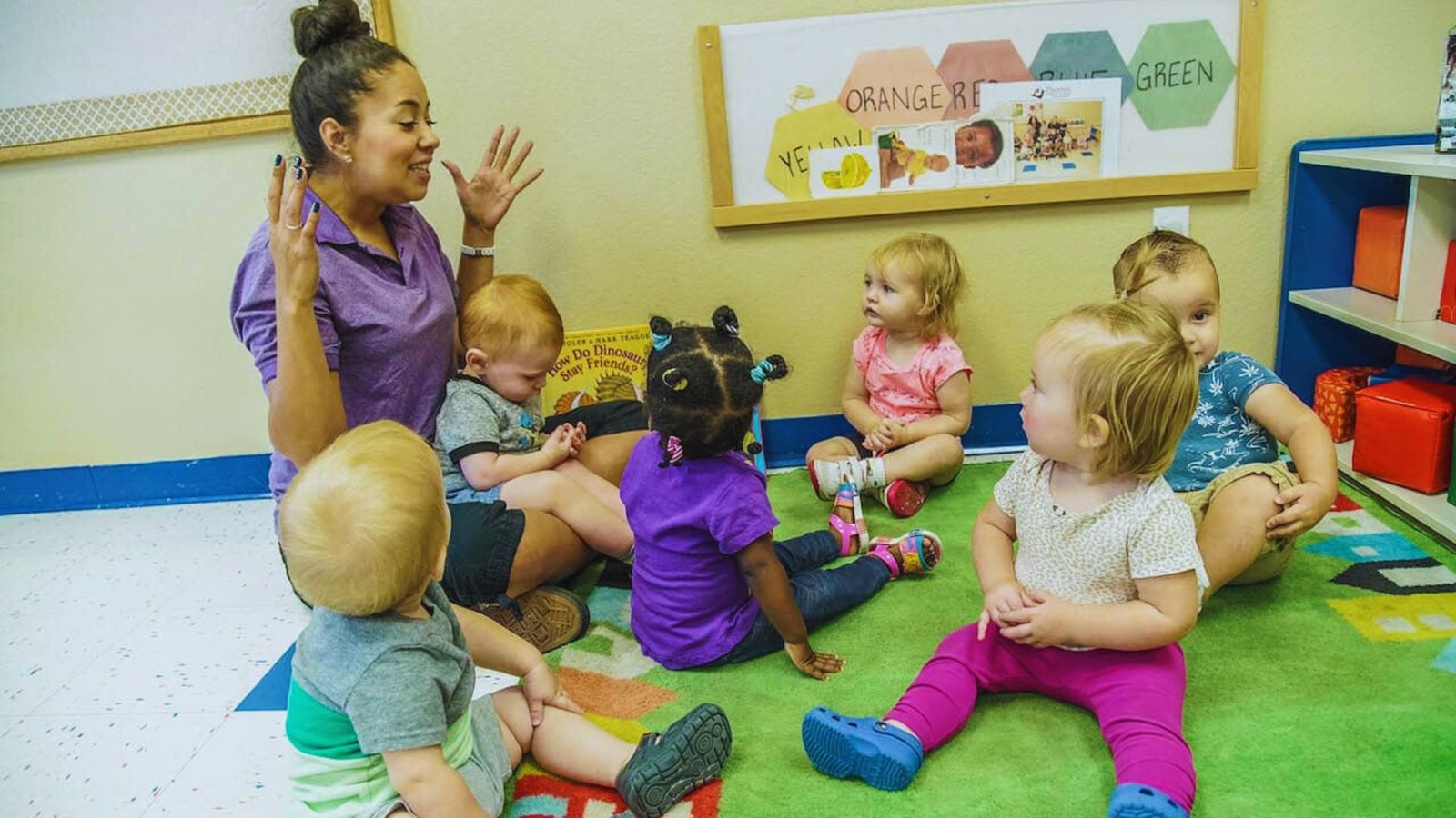 A woman sits in a circle with toddlers, engaging in activities during a daycare session for two-year-olds.
