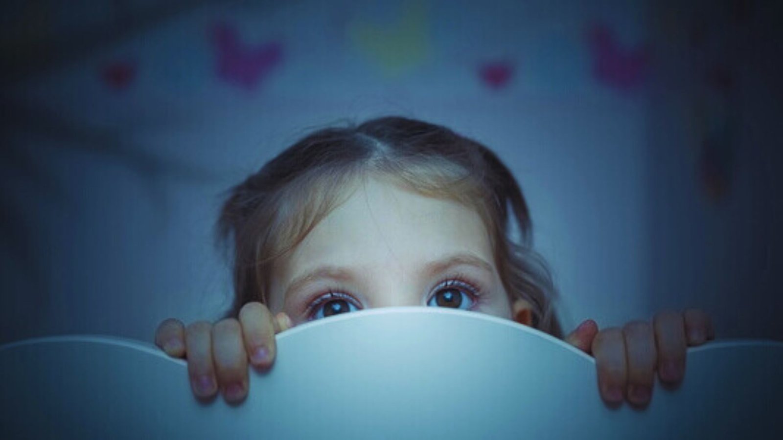 A young girl peeks out from behind a white wall, showing curiosity and a hint of fear, possibly related to being scared of the dark.
