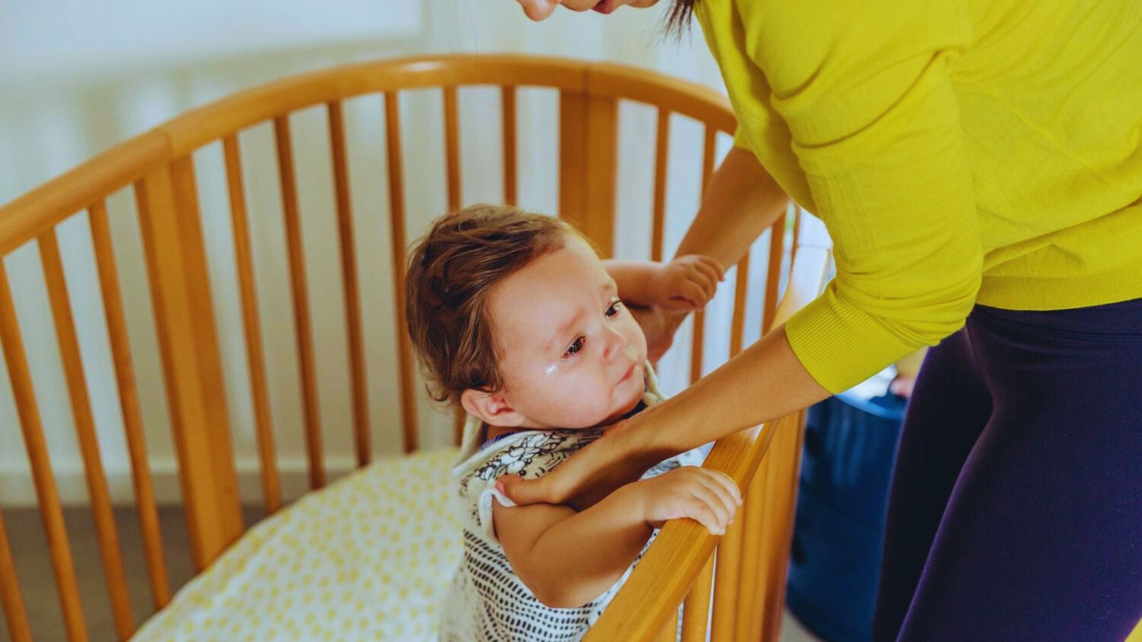 A woman gently assists a baby in a crib, illustrating parental support during the 2 to 1 nap transition.
